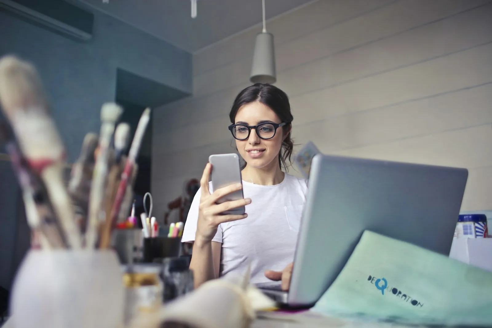 A woman at a work desk checking her smartphone next to a laptop