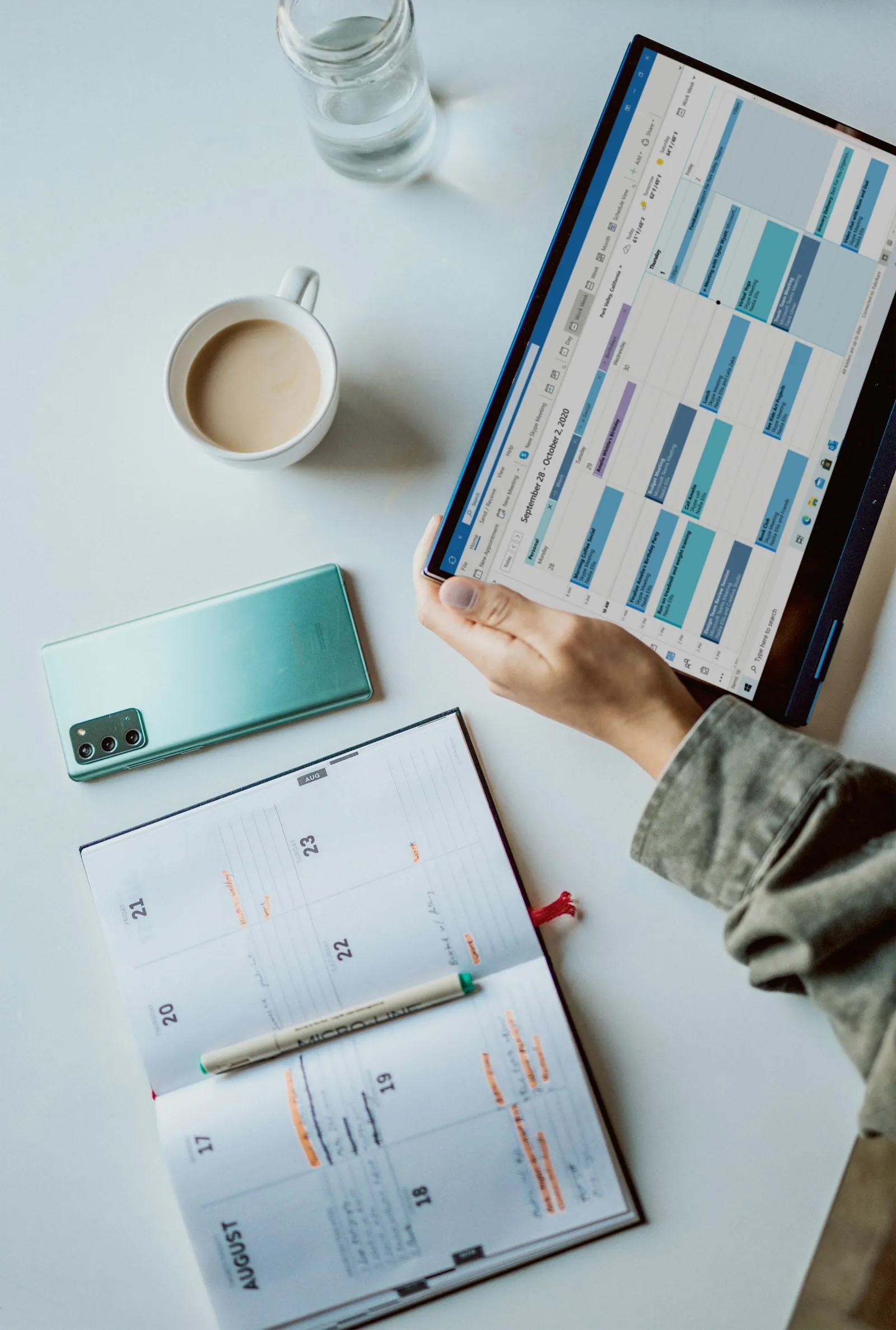 A hand holding a tablet with a weekly calendar, next to a paper planner, a phone and a cup of coffee