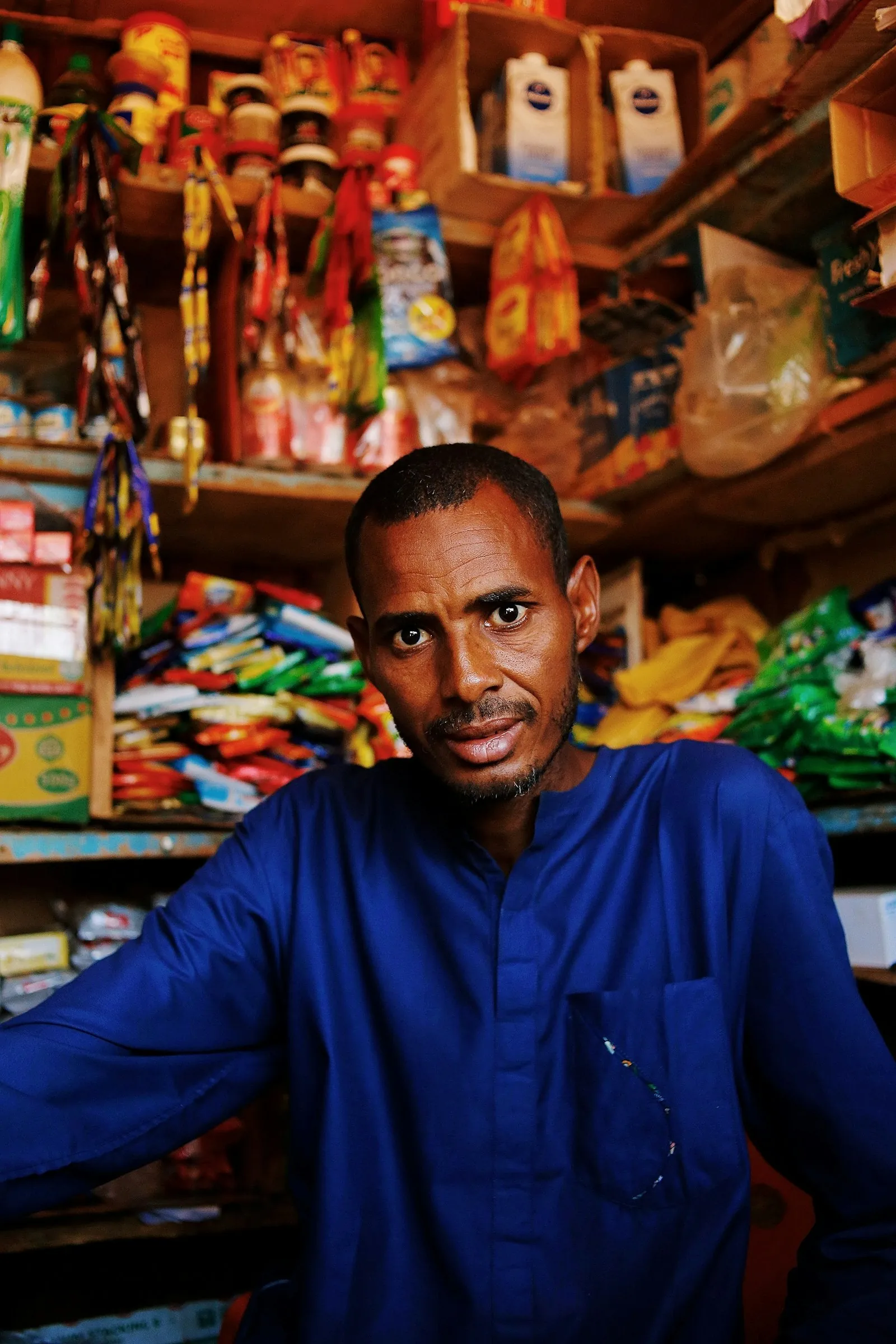 A shop owner behind the counter of his small store, with shelves of goods behind him