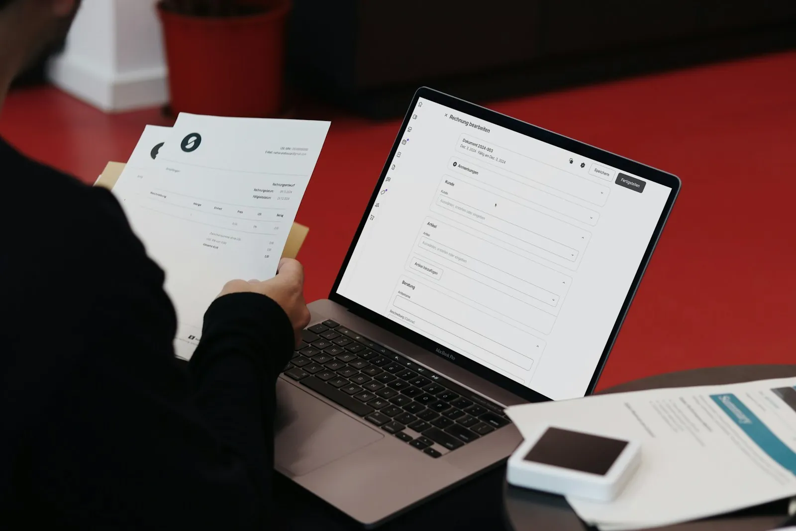 A person holding printed invoices while editing an invoice on a laptop