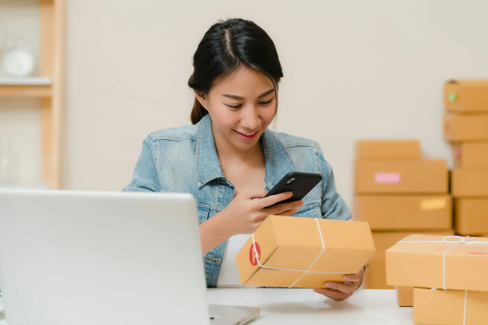 A small business owner checking a package with her phone next to a laptop and boxes